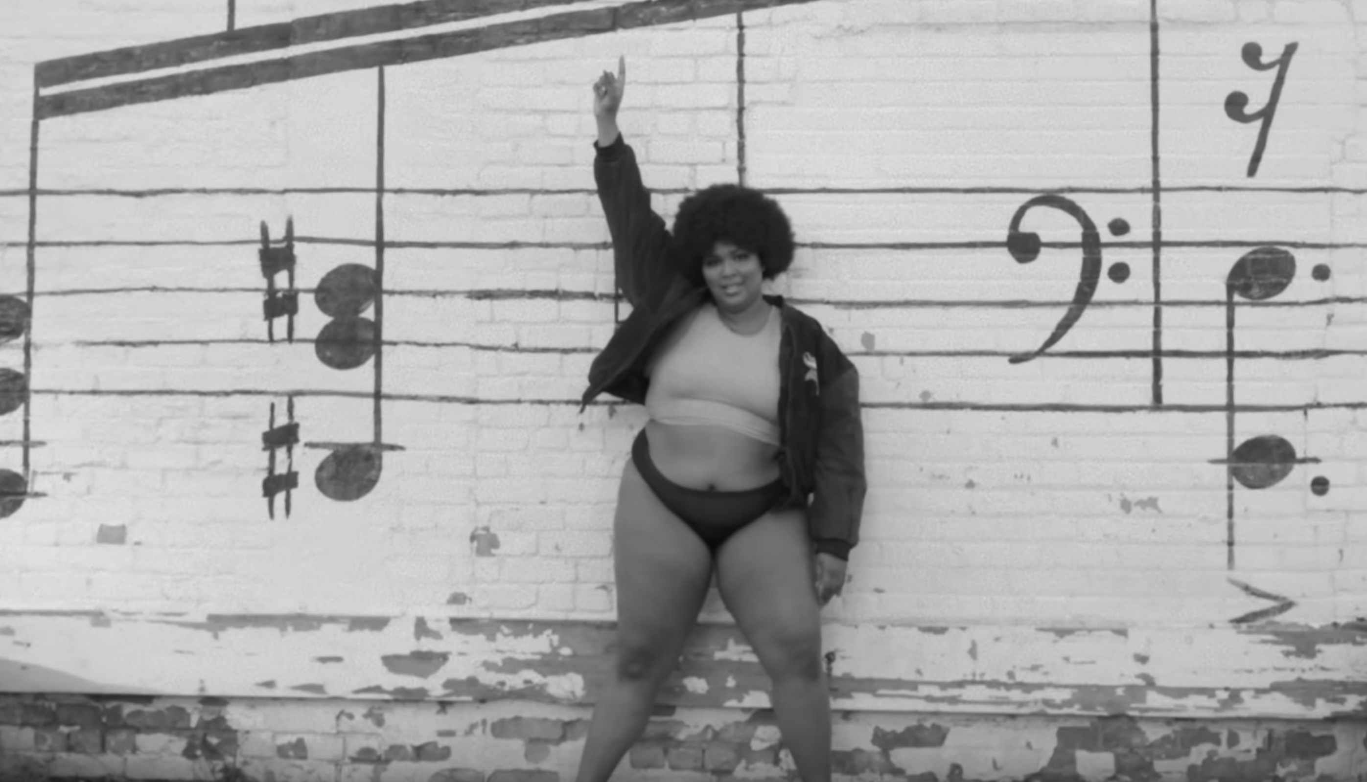 Greyscale photo of a woman (Lizzo) striking a magnificent pose in front of a brick wall with musical notation painted on it